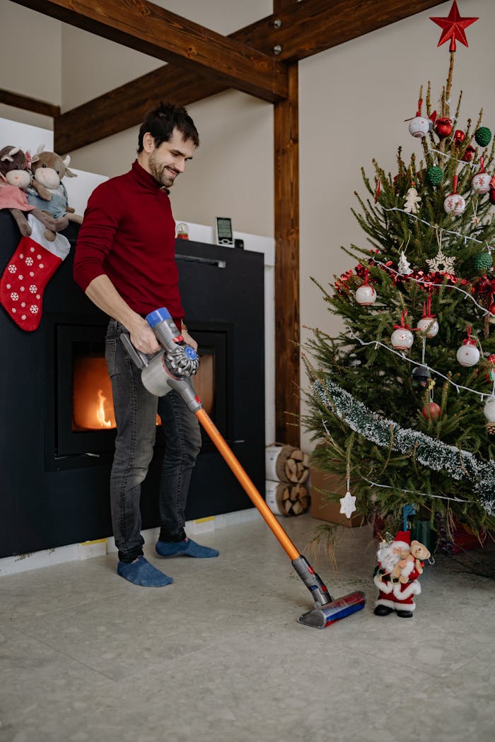 A man vacuuming the floor beside a decorated Christmas tree with holiday decor and a fireplace.
